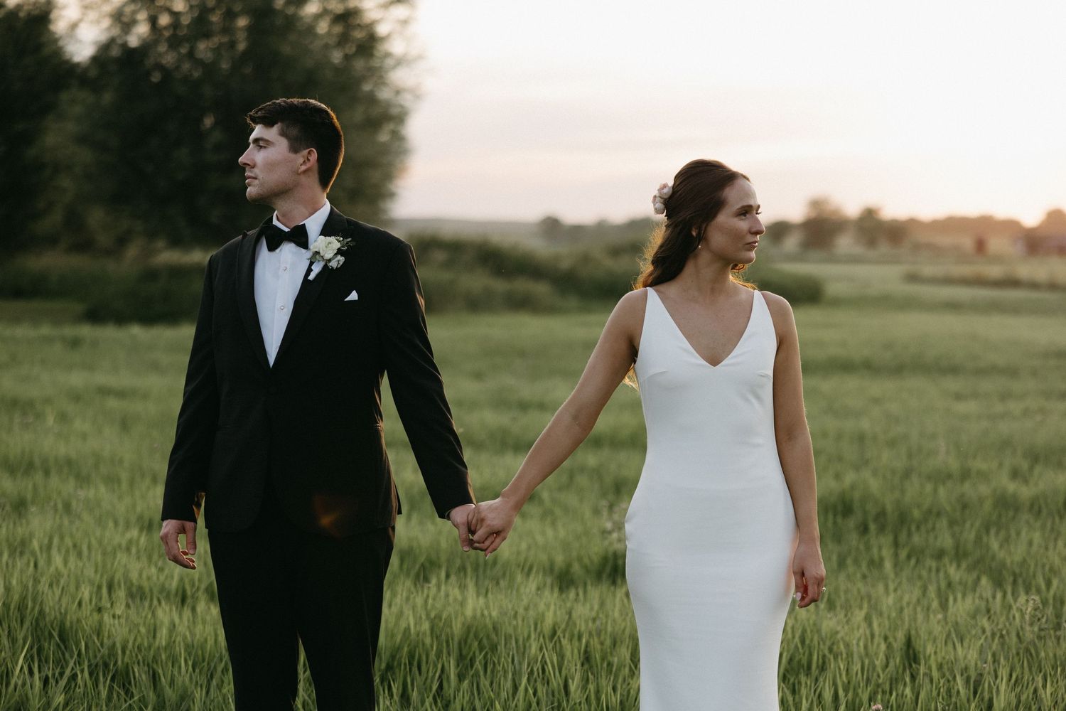 Newlyweds hold hands while walking through a lush green field at sunset wearing classic wedding attire.