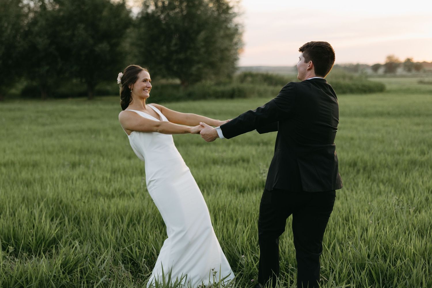 Wedding couple playfully dances in an open field during golden hour wearing formal attire.