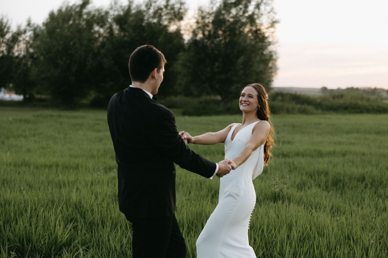 Couple in wedding attire dances and twirls in a verdant meadow at dusk.