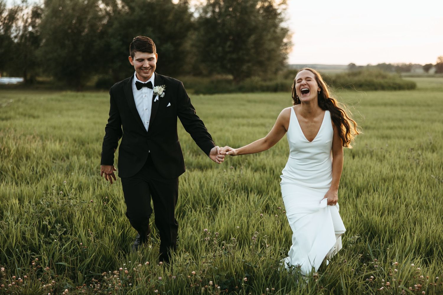 Wedding couple runs joyfully through tall grass field during sunset photoshoot.