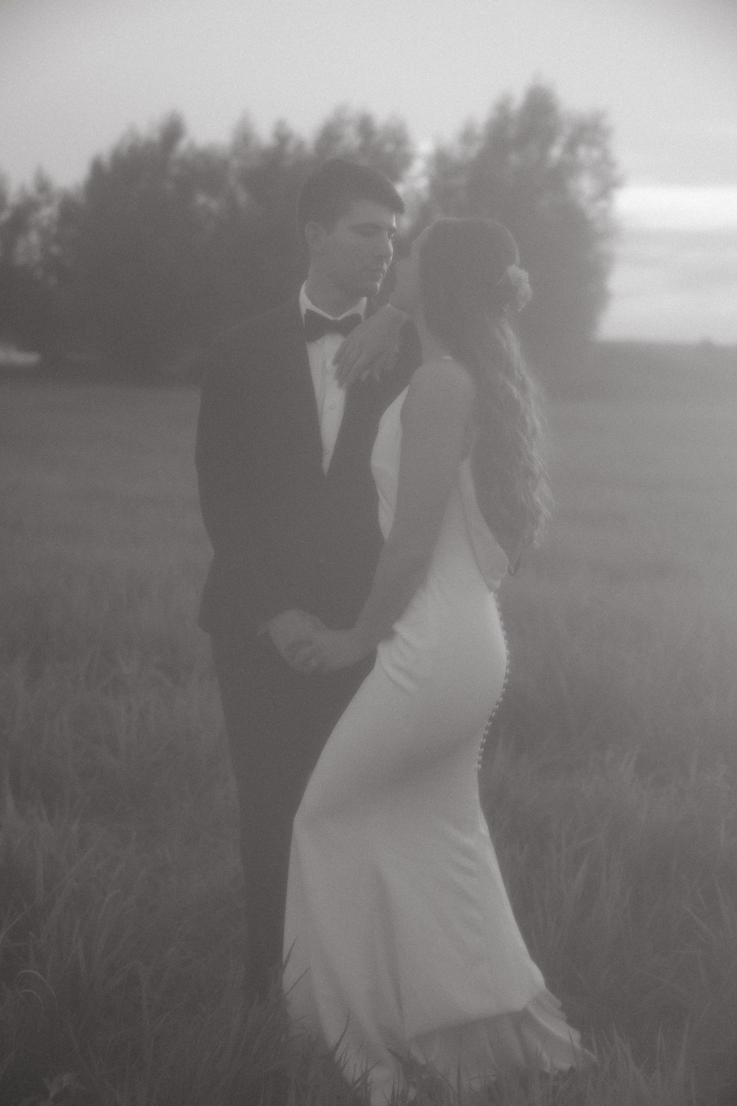 Atmospheric black and white photo of couple in wedding attire standing in misty field.