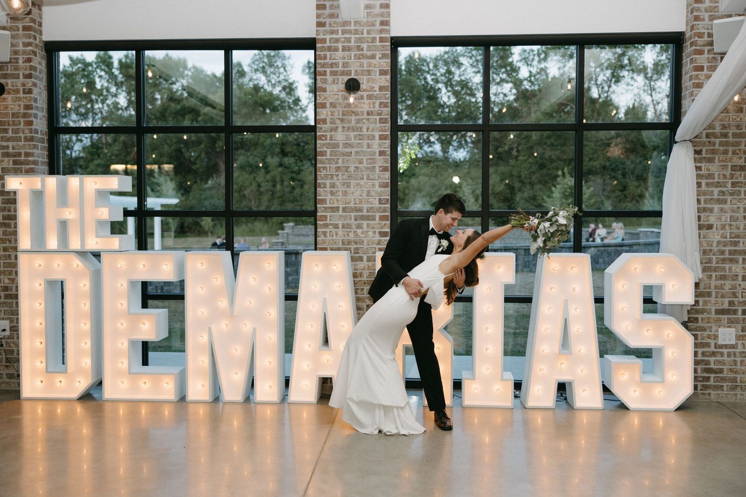 Couple shares romantic dance pose in front of large illuminated wedding name sign.