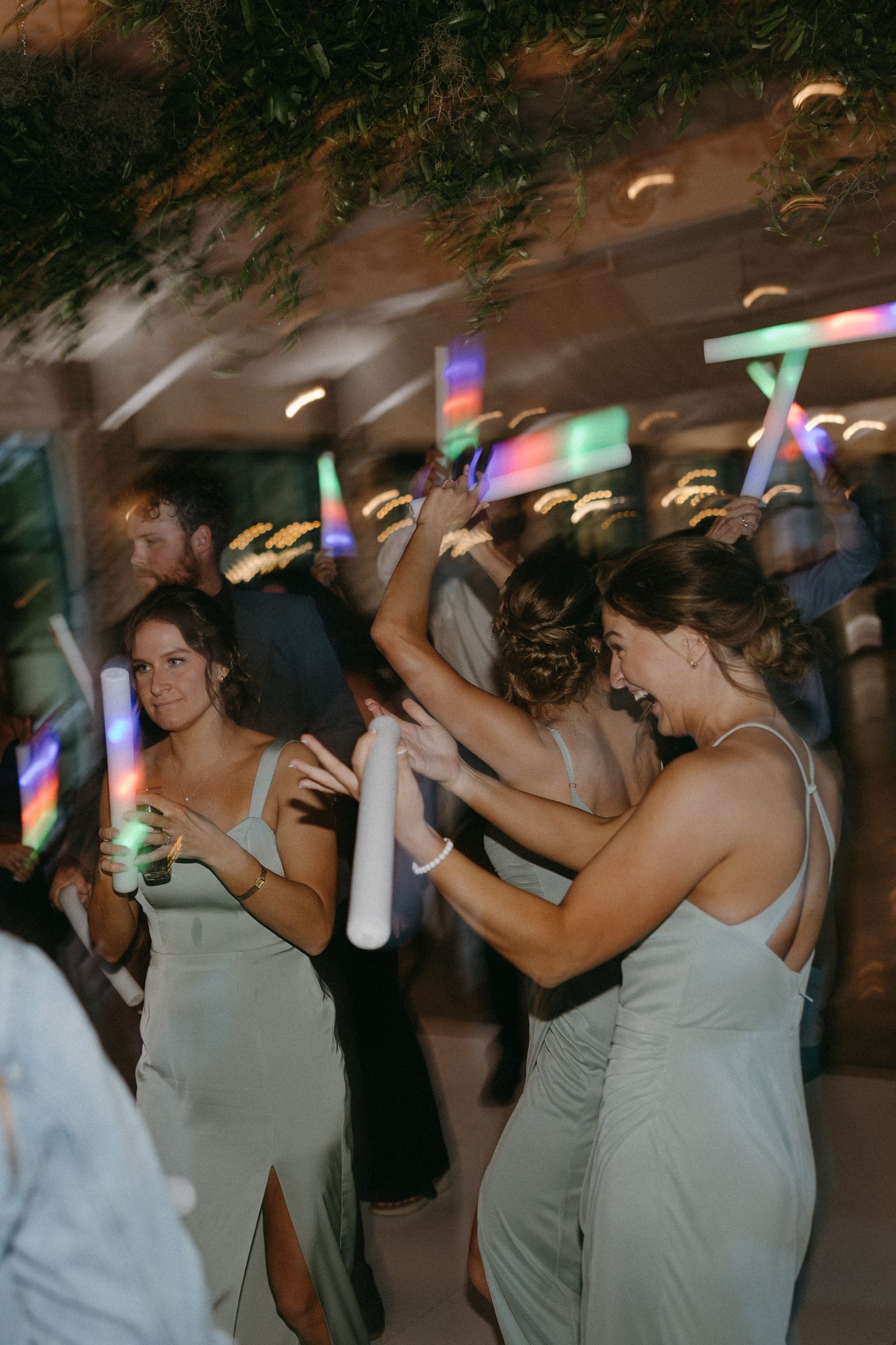 Wedding reception dance party with people holding colorful light sticks under dim lighting.