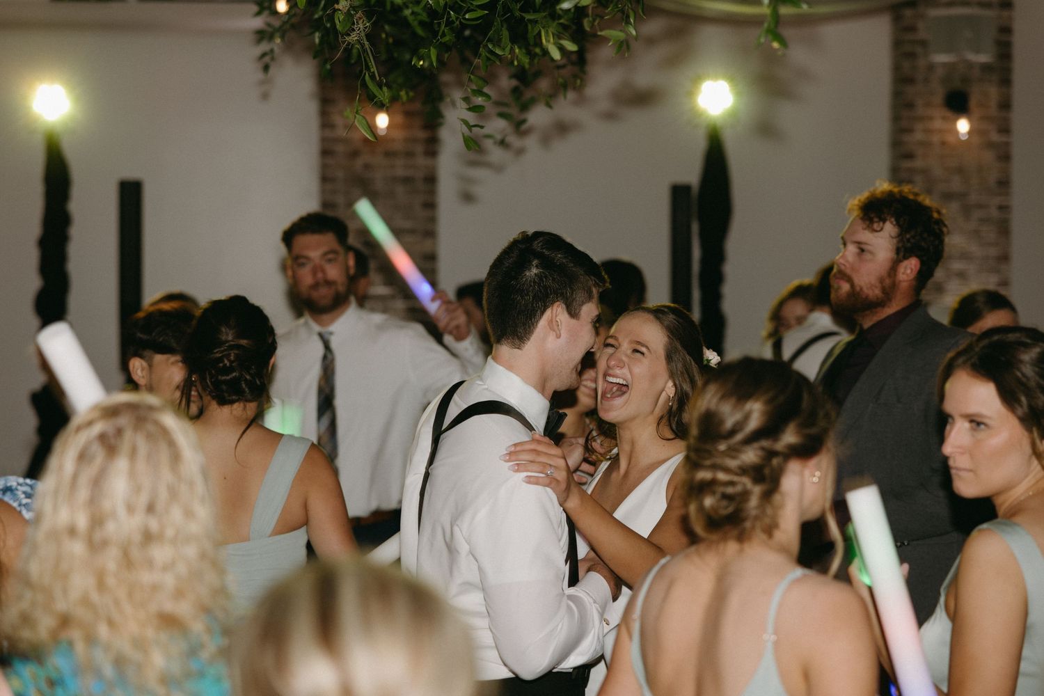 Lively wedding reception scene with guests dancing and holding light-up wands under greenery decor.