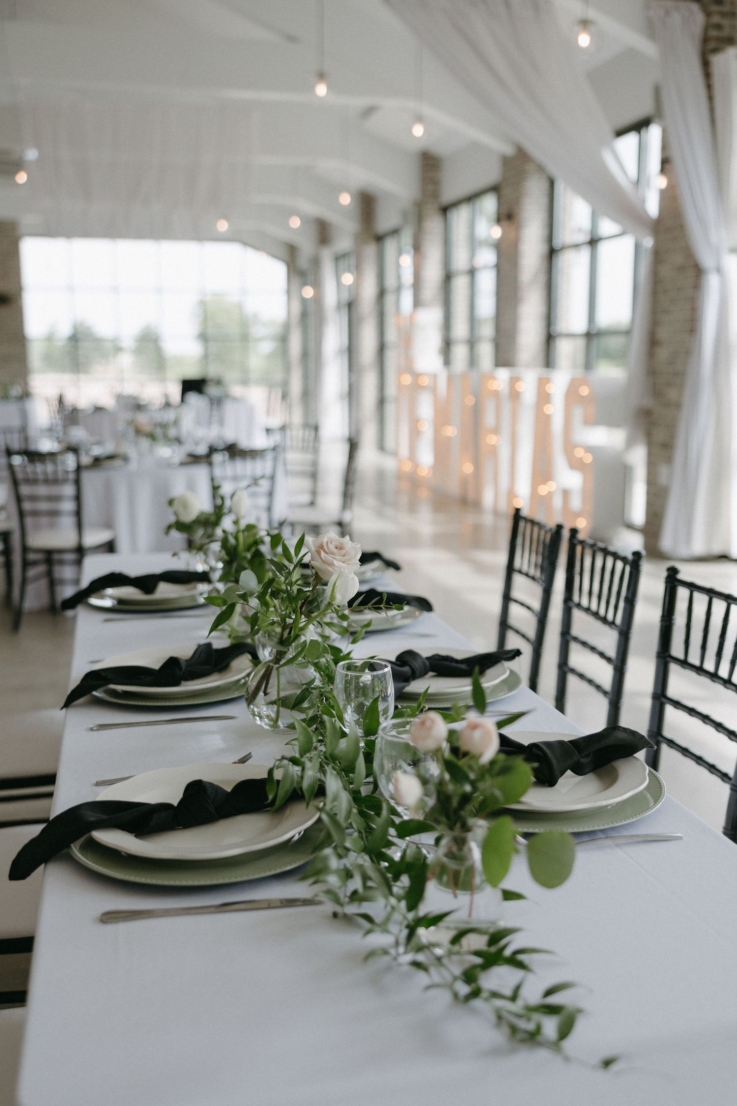Elegant wedding reception table setup with white linens, black accents and natural greenery centerpieces in a bright venue.