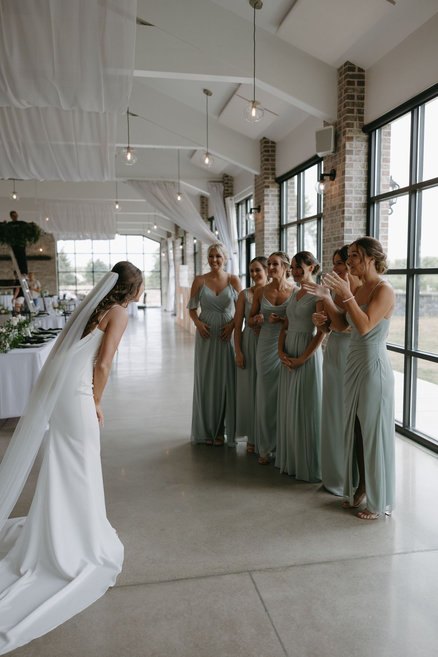 Bridesmaids in sage green dresses react emotionally to seeing bride in white gown in bright venue hallway.