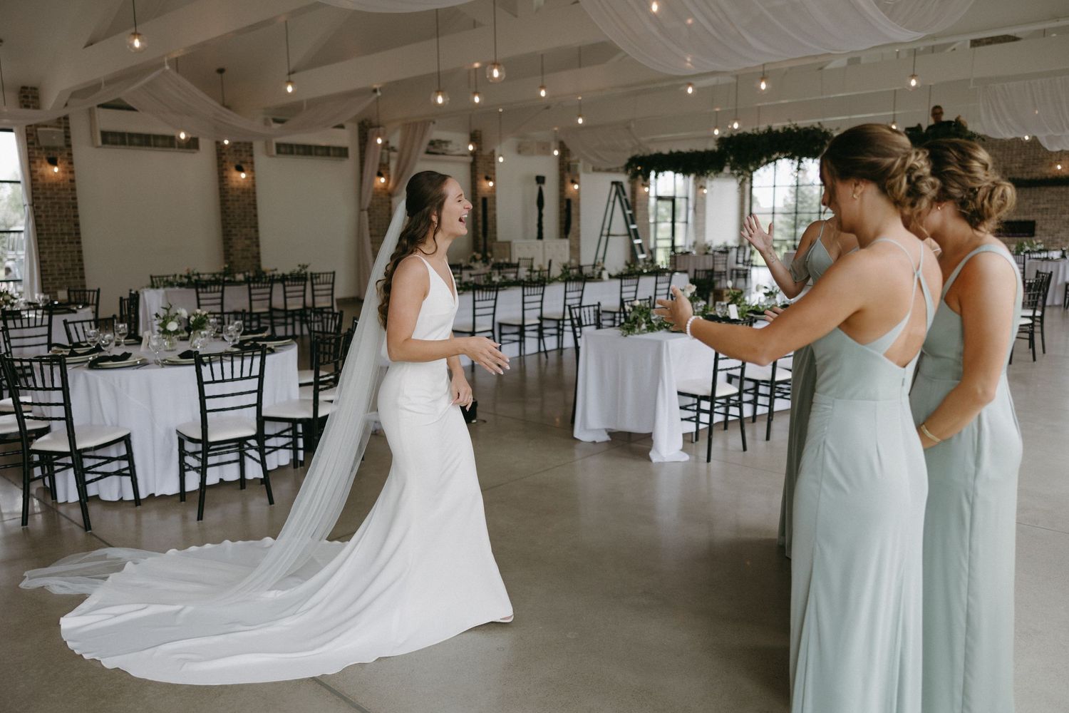 Bride shows off wedding dress train while bridesmaids in mint dresses admire in modern reception venue.