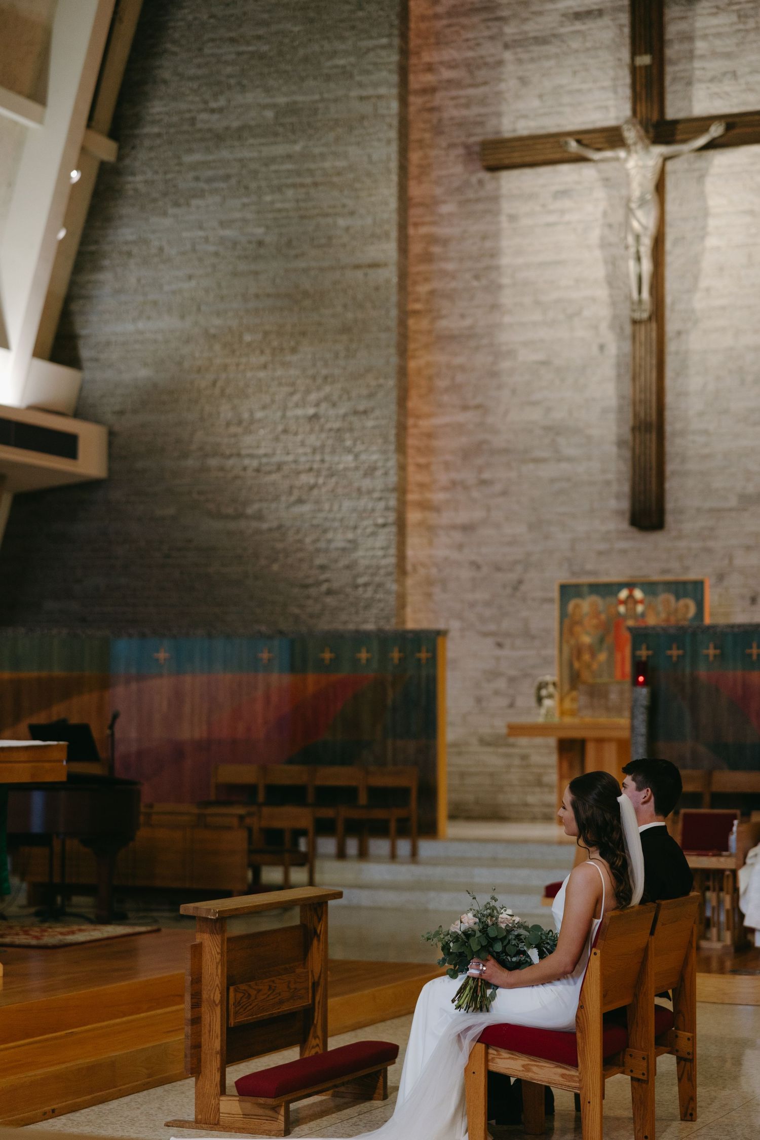 Wedding ceremony in church interior with wooden pews and large crucifix on brick wall.