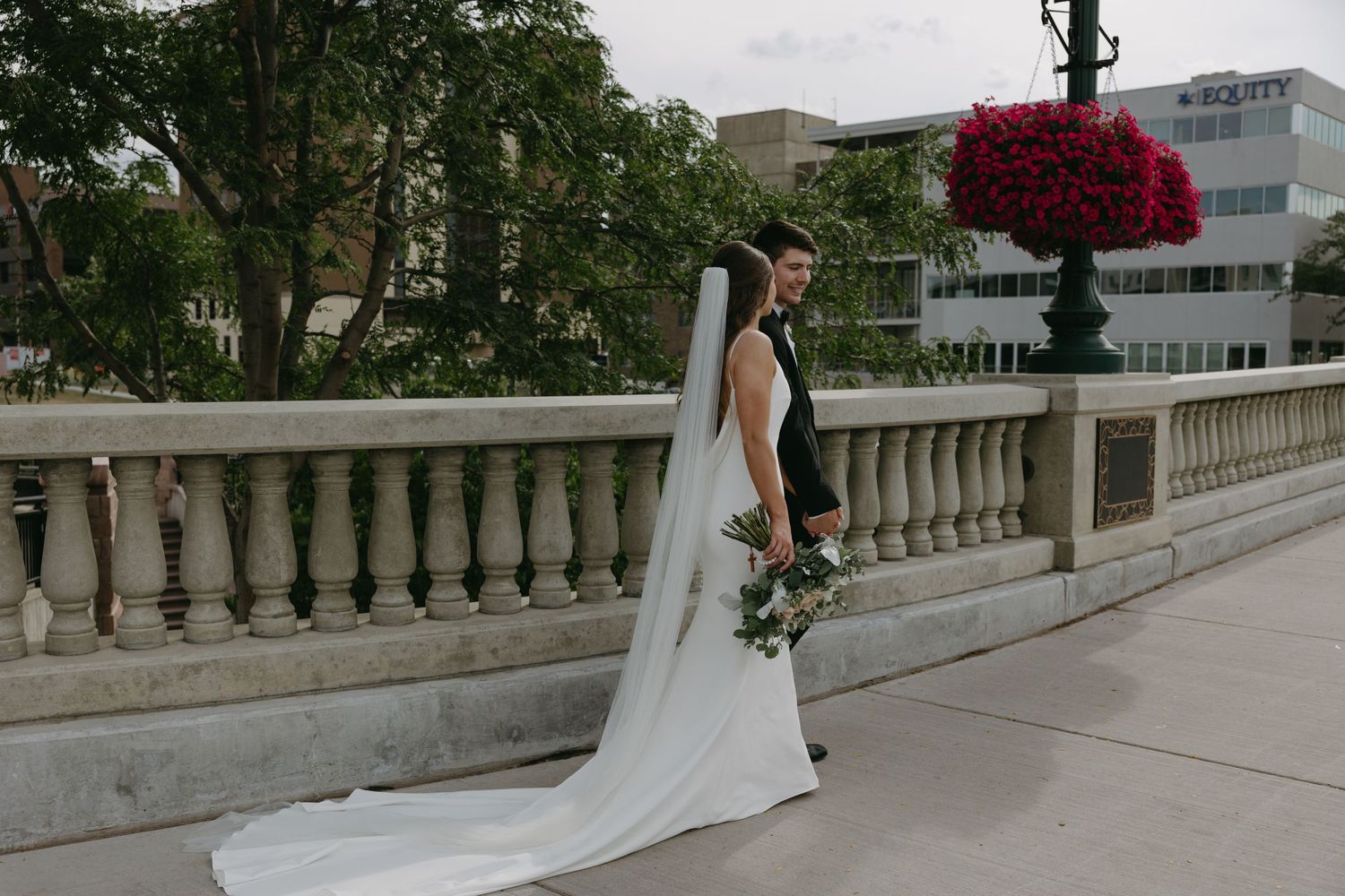 Bride in sleek white gown poses on stone balcony with flowing veil and bouquet.