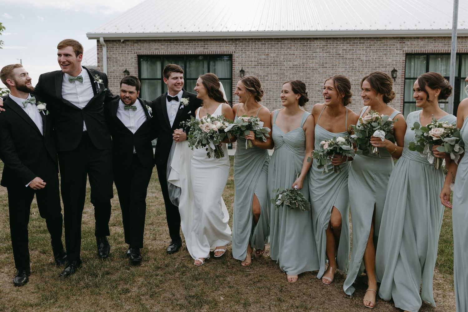 Wedding party in formal attire poses outdoors with bridesmaids wearing light blue dresses and holding bouquets.