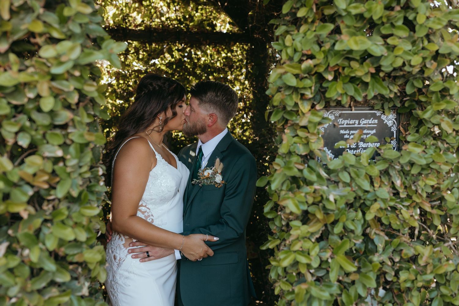 couple in wedding attire kissing while standing between leafy hedges in an outdoor secret garden setting.