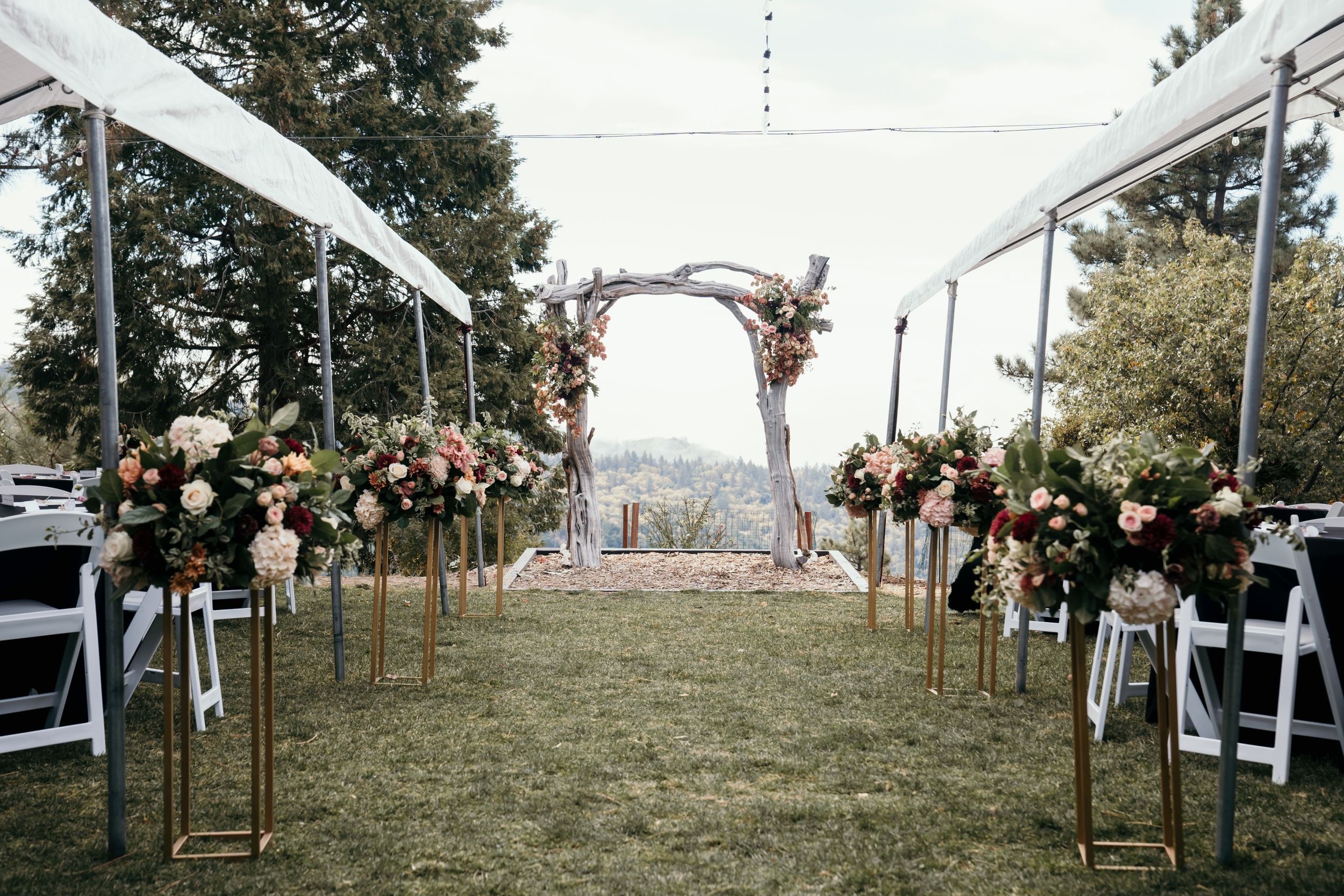 Outdoor wedding ceremony setup with floral arch and white chairs on grassy hillside.