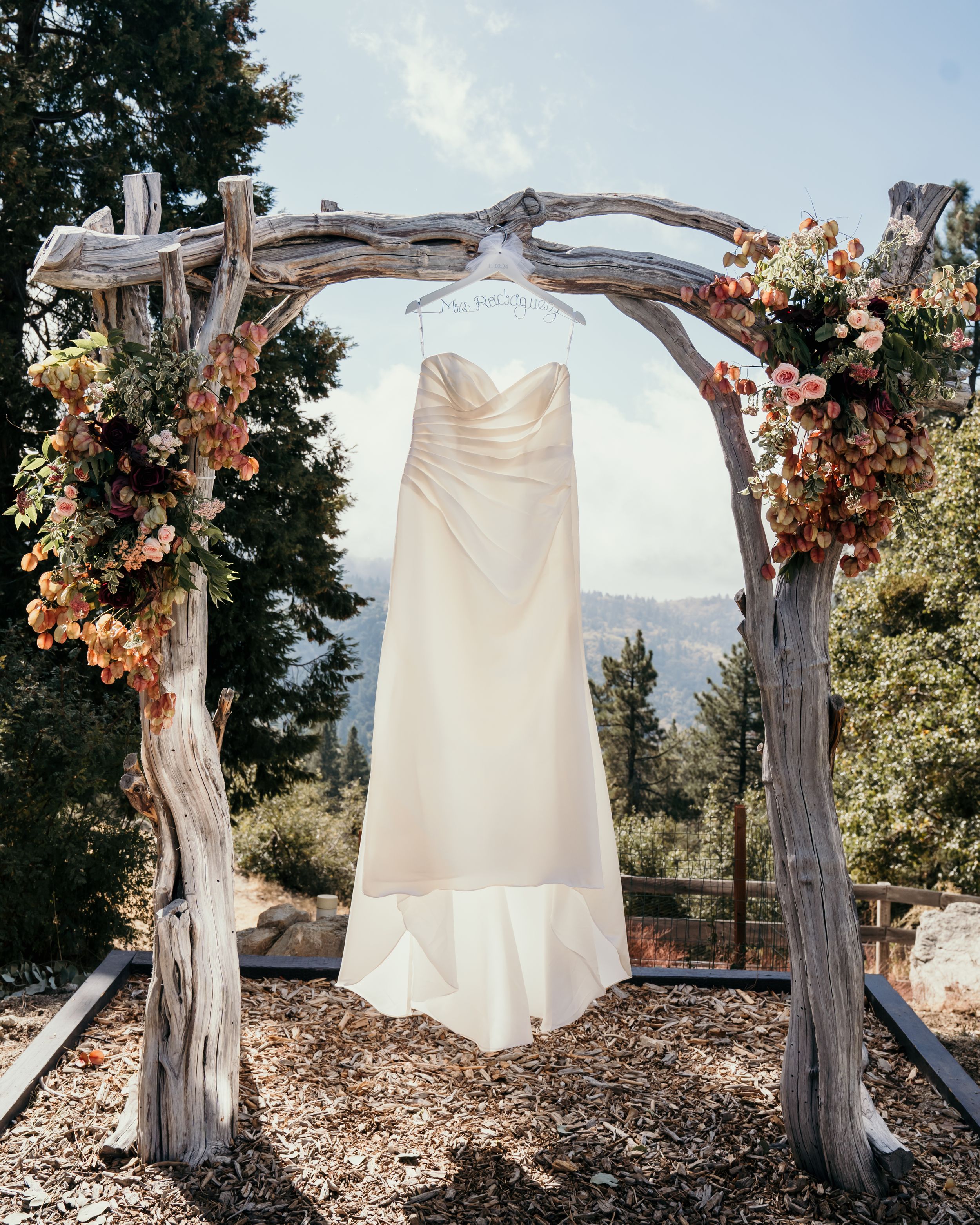 White wedding dress hanging from rustic driftwood arch decorated with peach and pink flowers.