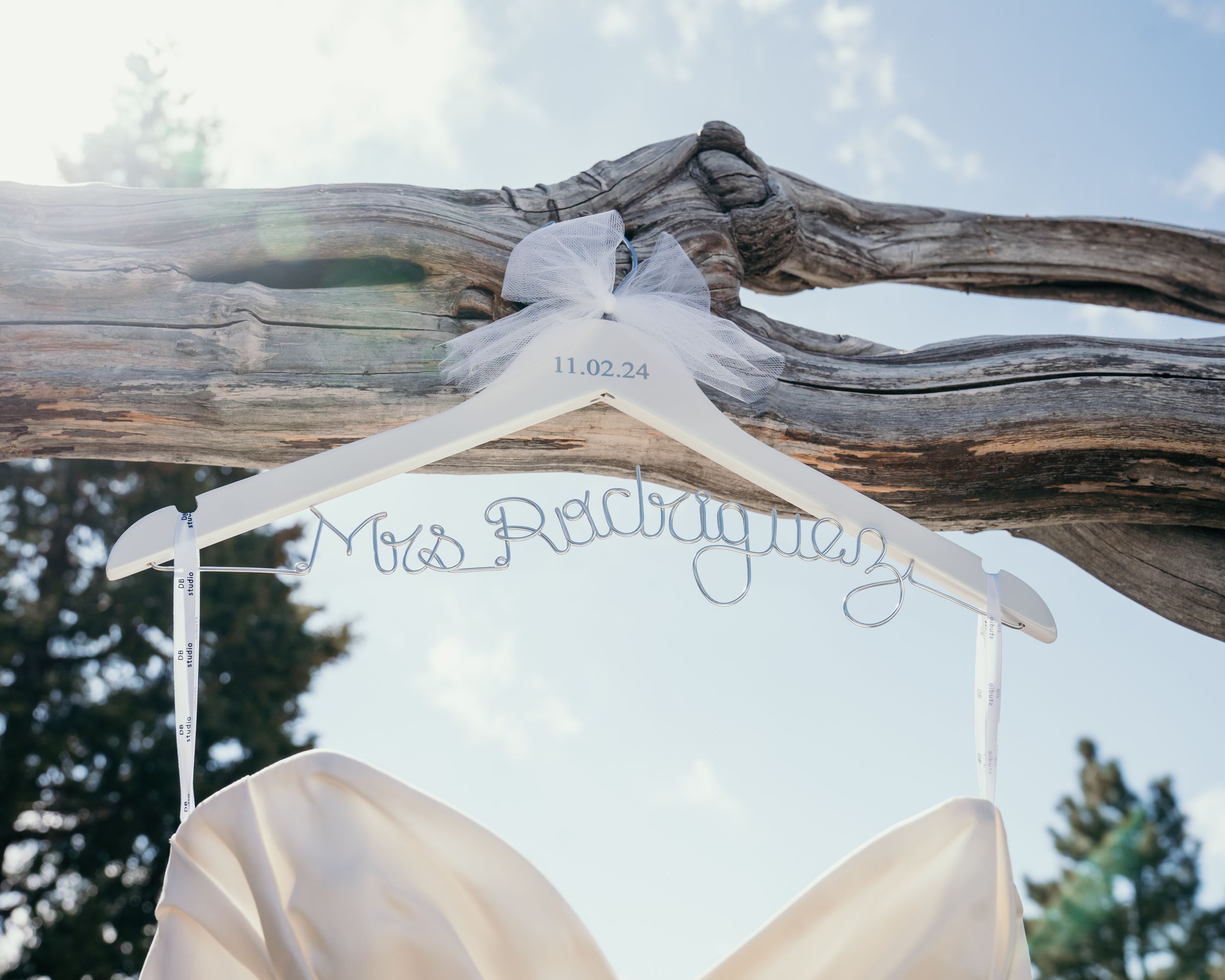 Wedding dress hanging on personalized bridal hanger against bright sky.