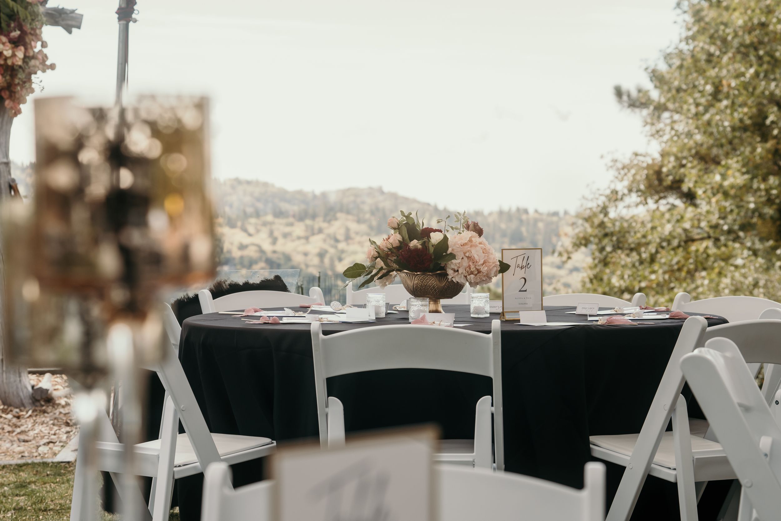 Outdoor wedding reception table setup with black chairs and mountain view backdrop.