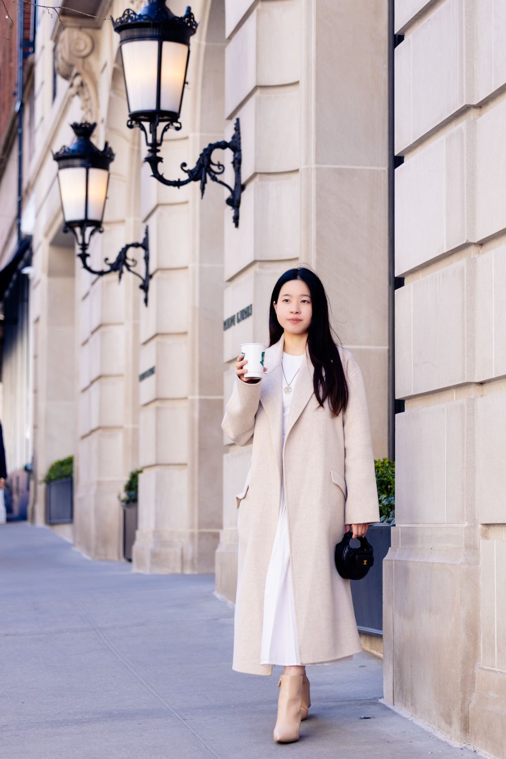 Elegant street fashion photo showing a person in a beige coat and white dress holding coffee against classic architecture.