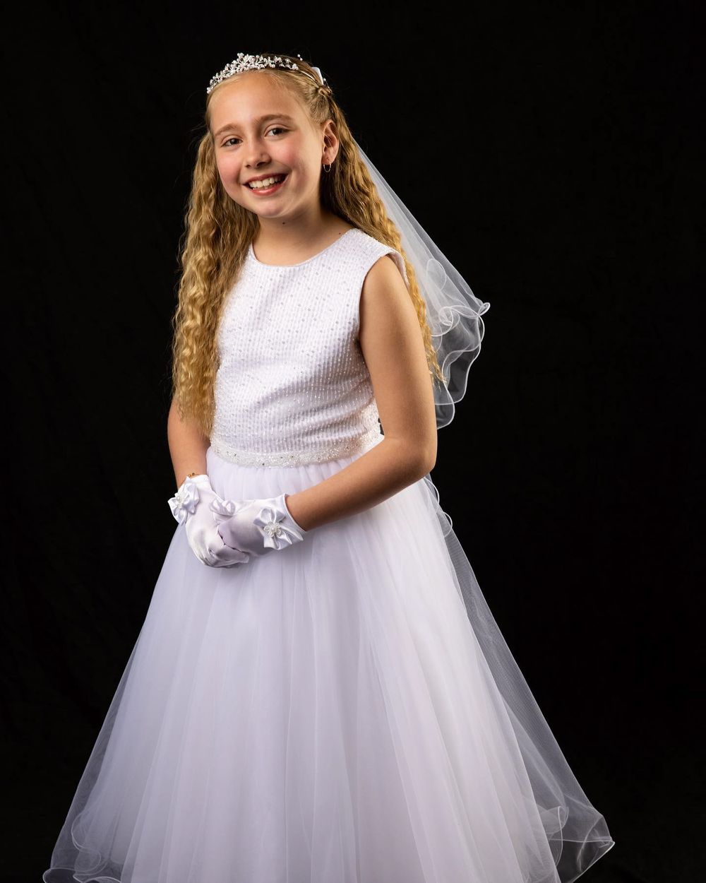First communion portrait of young person in white dress, veil and crown against dark background.