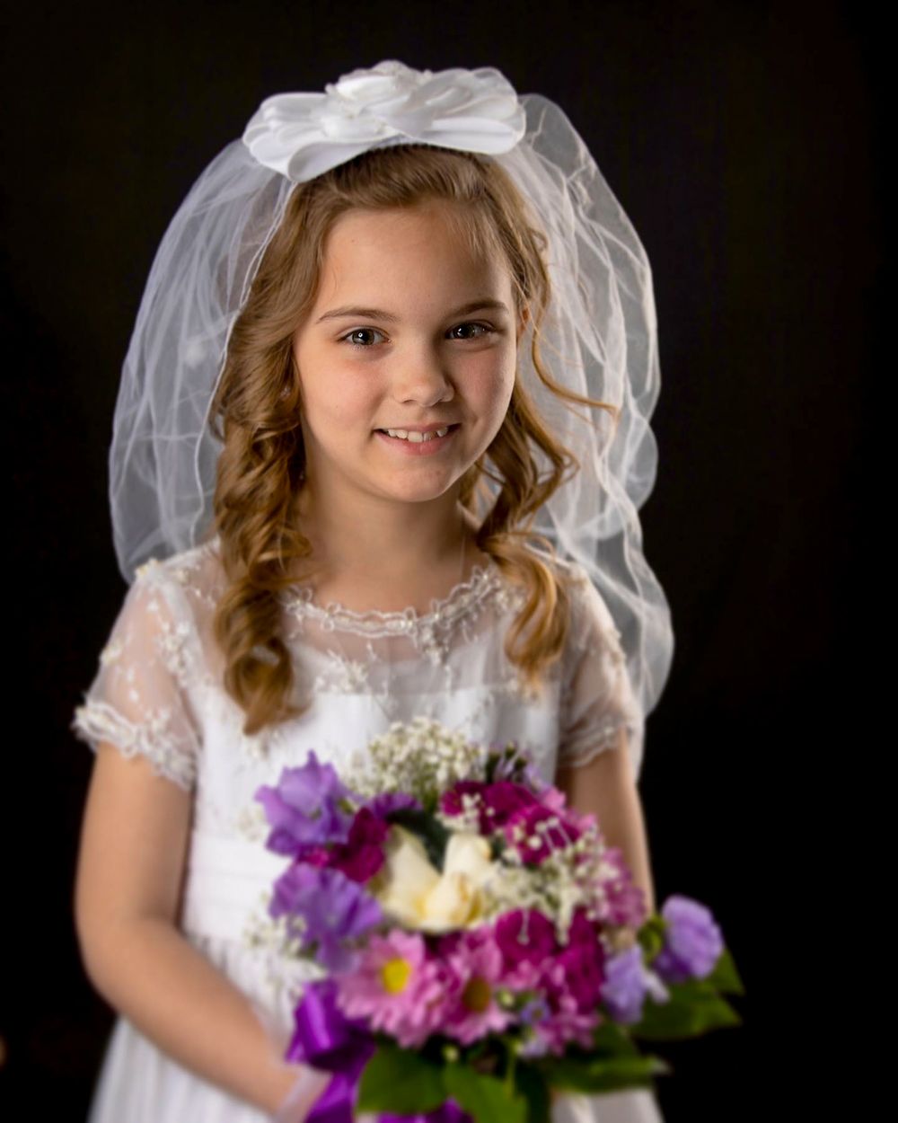 First communion portrait with colorful purple and white flower bouquet against dark backdrop.