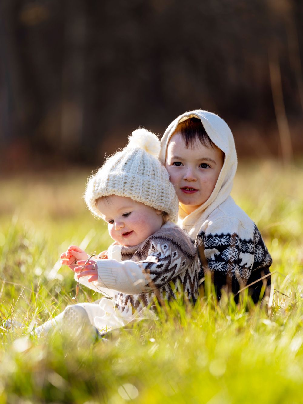 Two children sit together in sunlit grass wearing winter clothing on a bright autumn day.