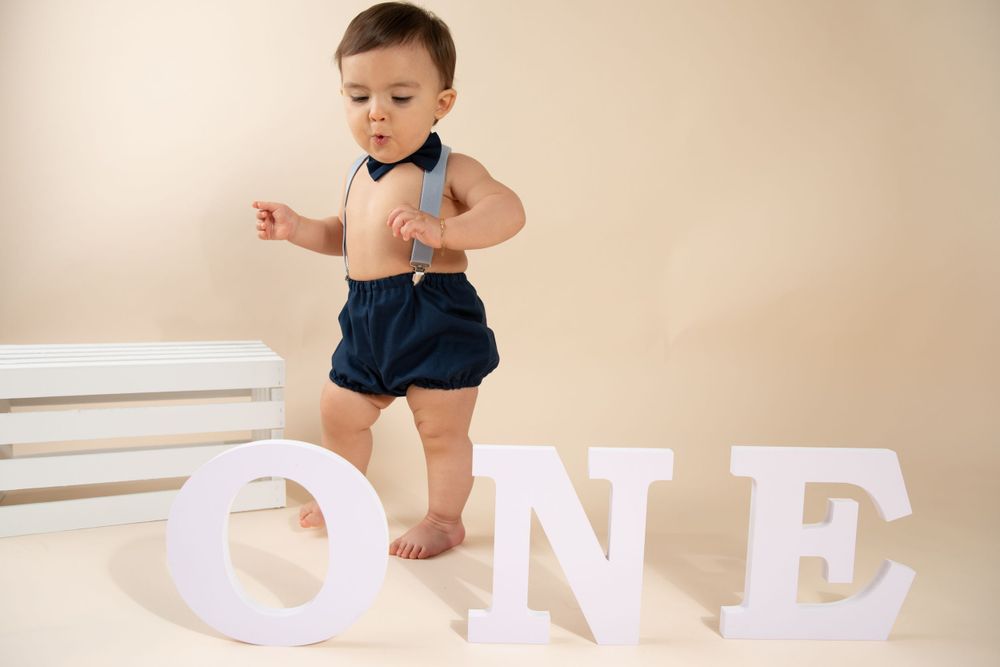 A toddler in navy shorts standing next to white 'ONE' letters against beige background.