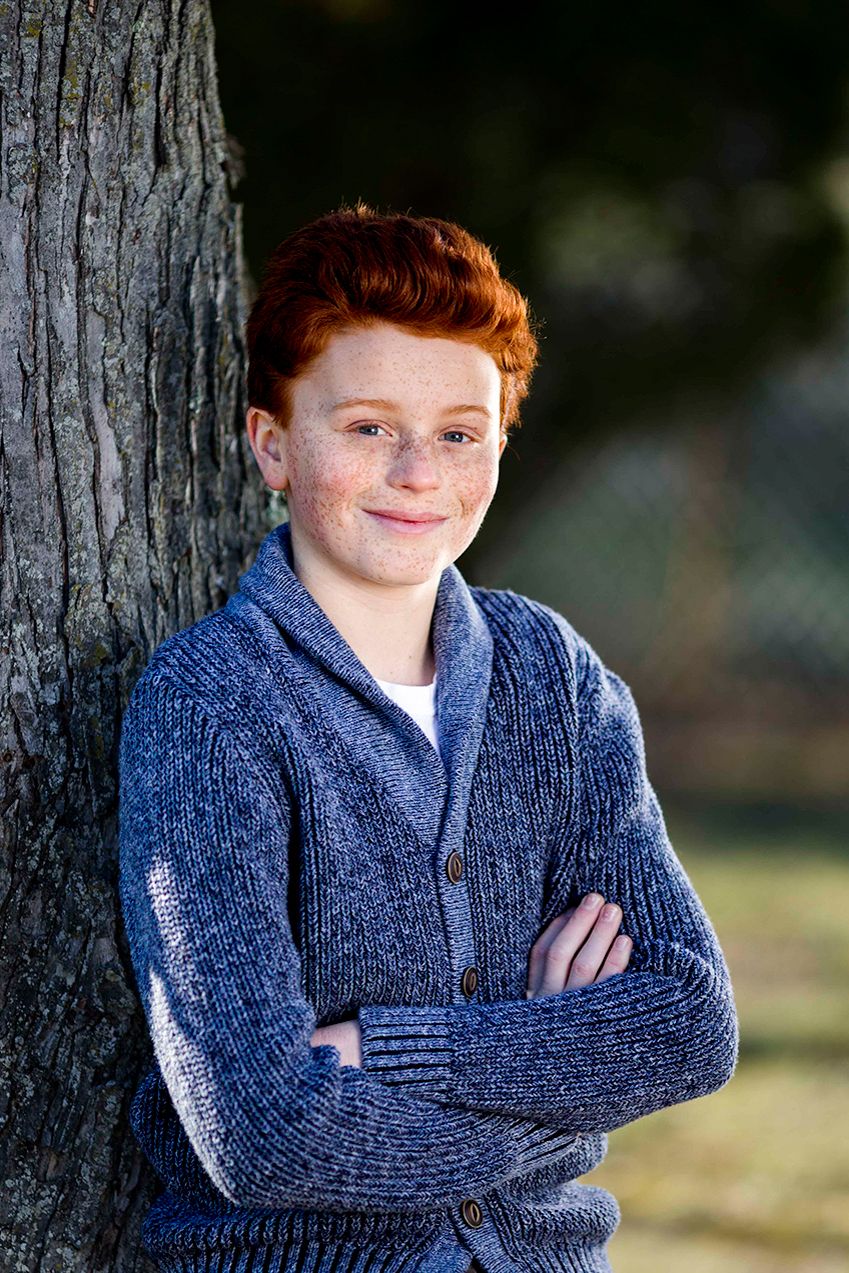 A portrait of a young person in a navy sweater leaning against a tree.