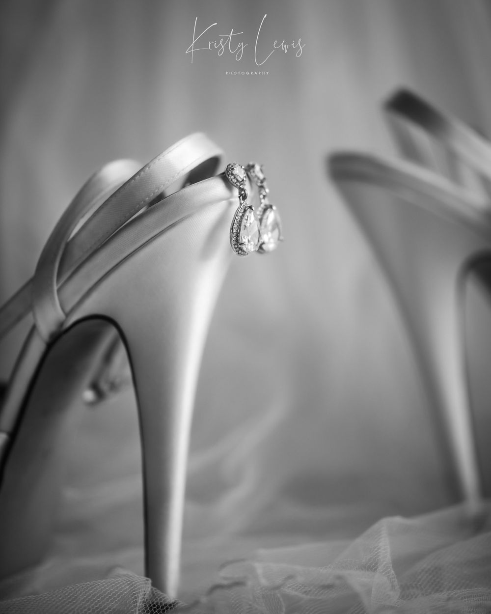 black and white product photo of white satin bridal shoes with diamond earrings hanging off the back of the shoe. Shot in Blackwood, Wales