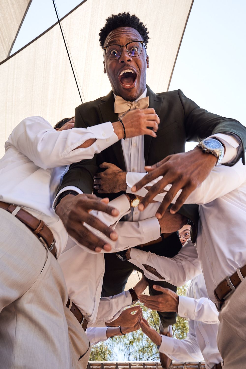 Wide low angle shot of groomsmen carrying groom and tossing him into the air as he looks excited before his wedding ceremony.