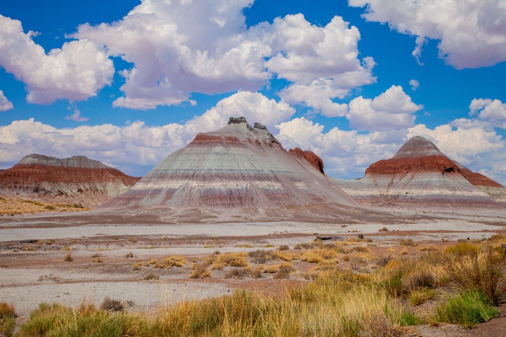 Colorful striped rock formations rise from desert landscape under blue sky with white puffy clouds.