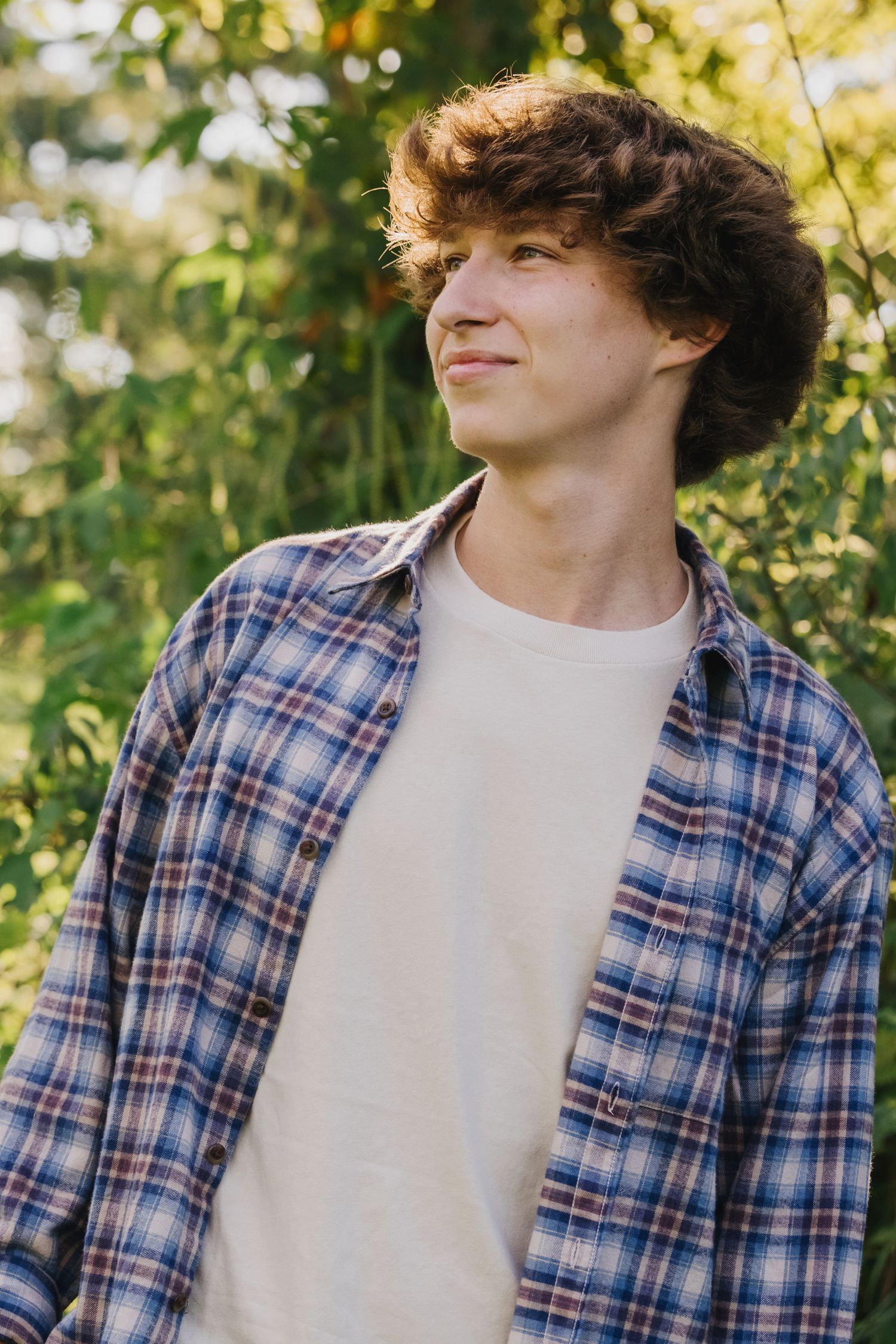 A natural portrait of someone wearing a plaid shirt and white tee against green foliage.
