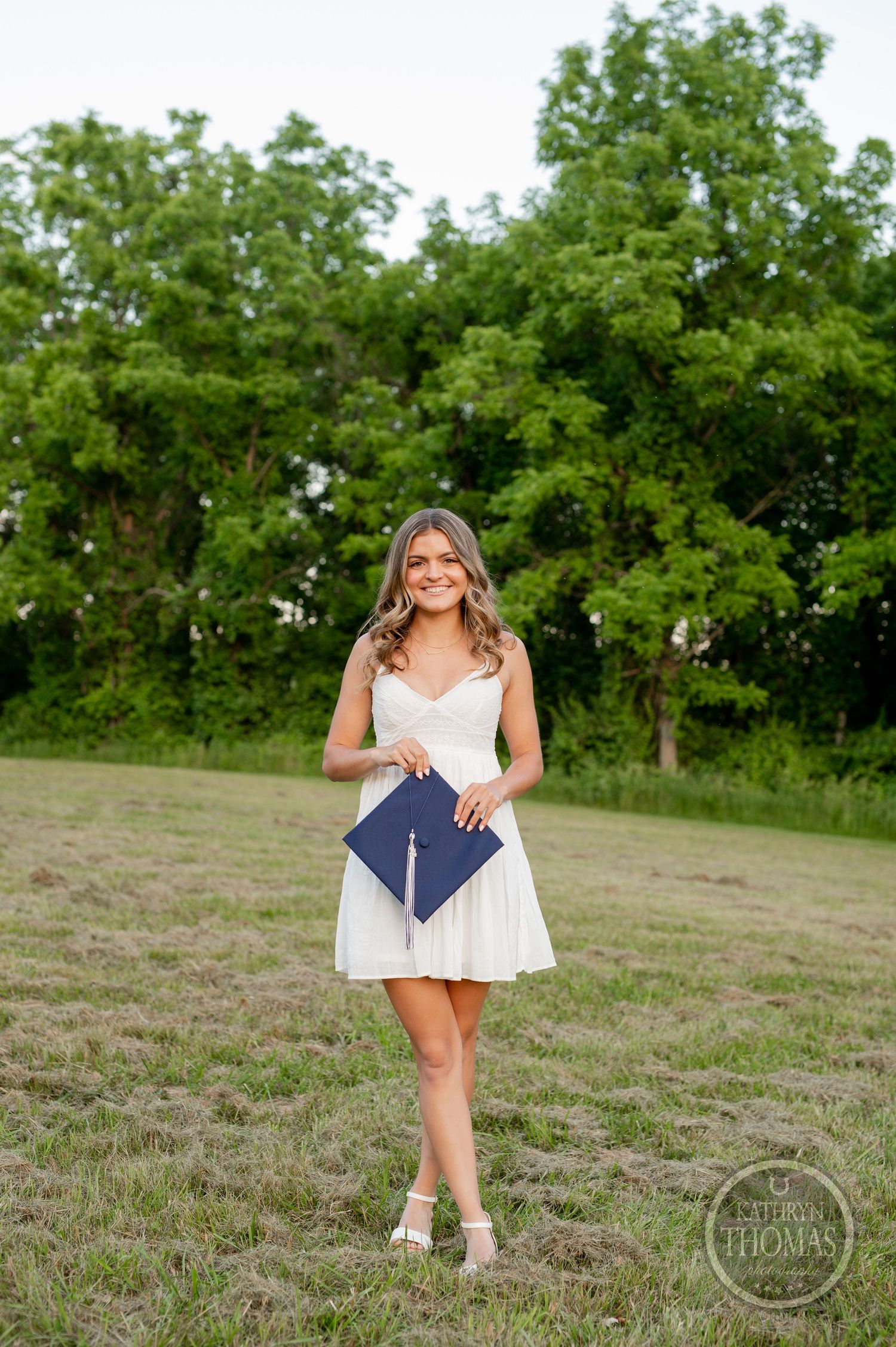 A graduate in white dress holds diploma while standing in a grassy field with trees.