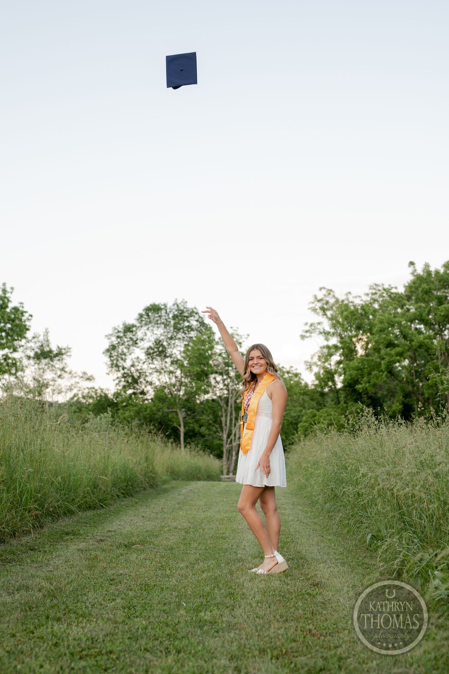 Graduate tosses cap in air while celebrating on grassy path during sunset.