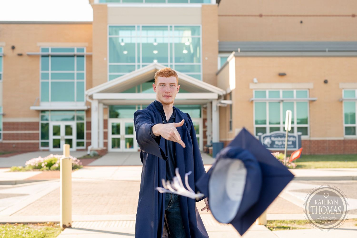 Graduate playfully tosses graduation cap in air outside academic building.