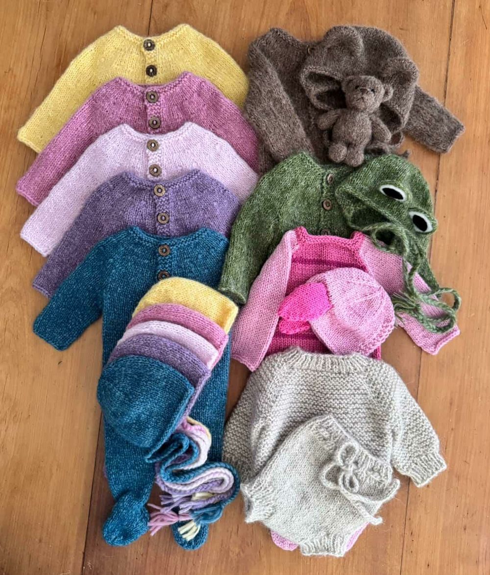 A collection of handmade knit baby clothes in soft pastel colors arranged on a wooden floor.