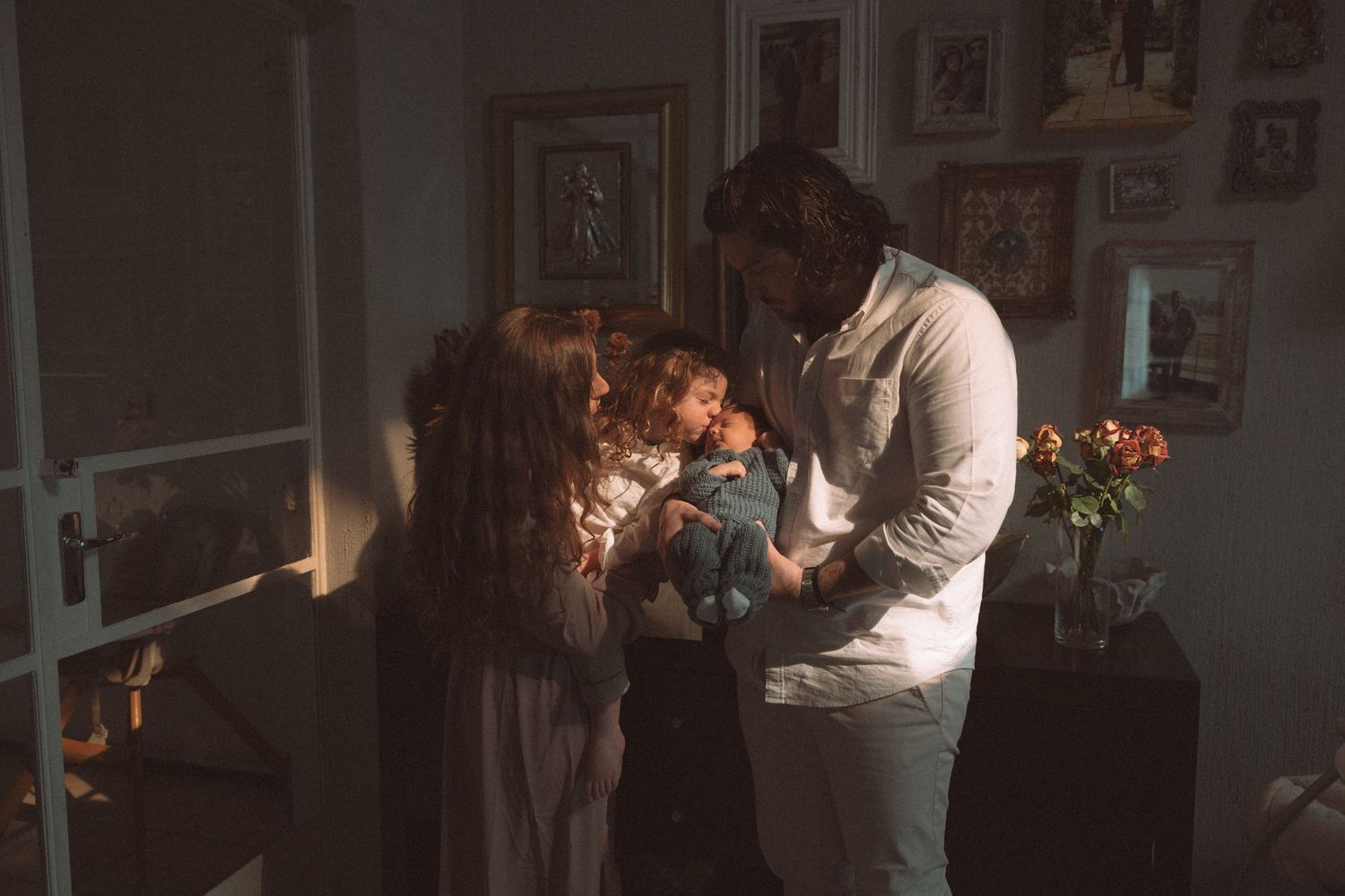 A family shares an intimate moment together in a dimly lit room with framed photos on the wall and flowers nearby.
