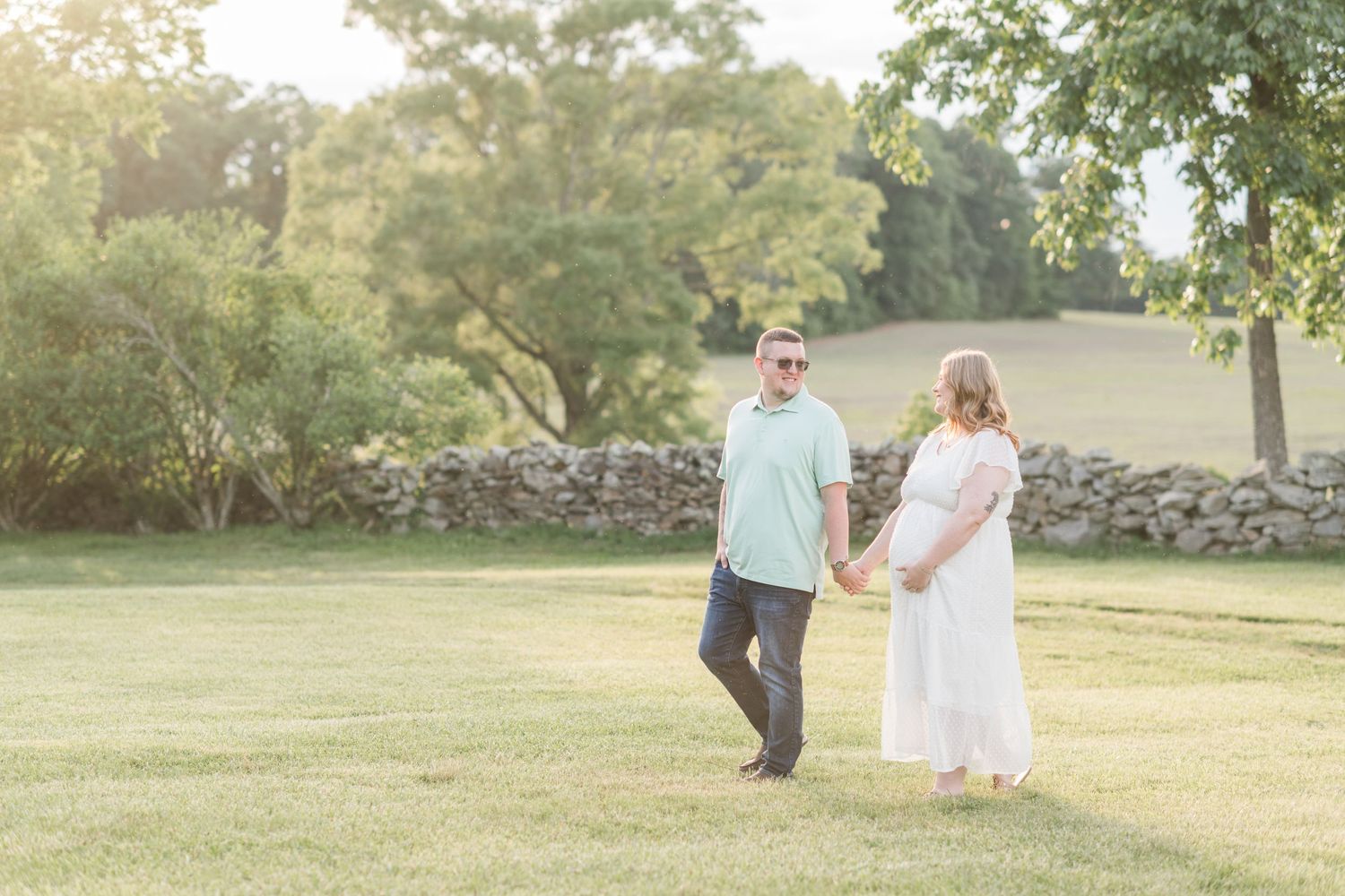 Pregnant couple holding hands walking through a sunlit green lawn.