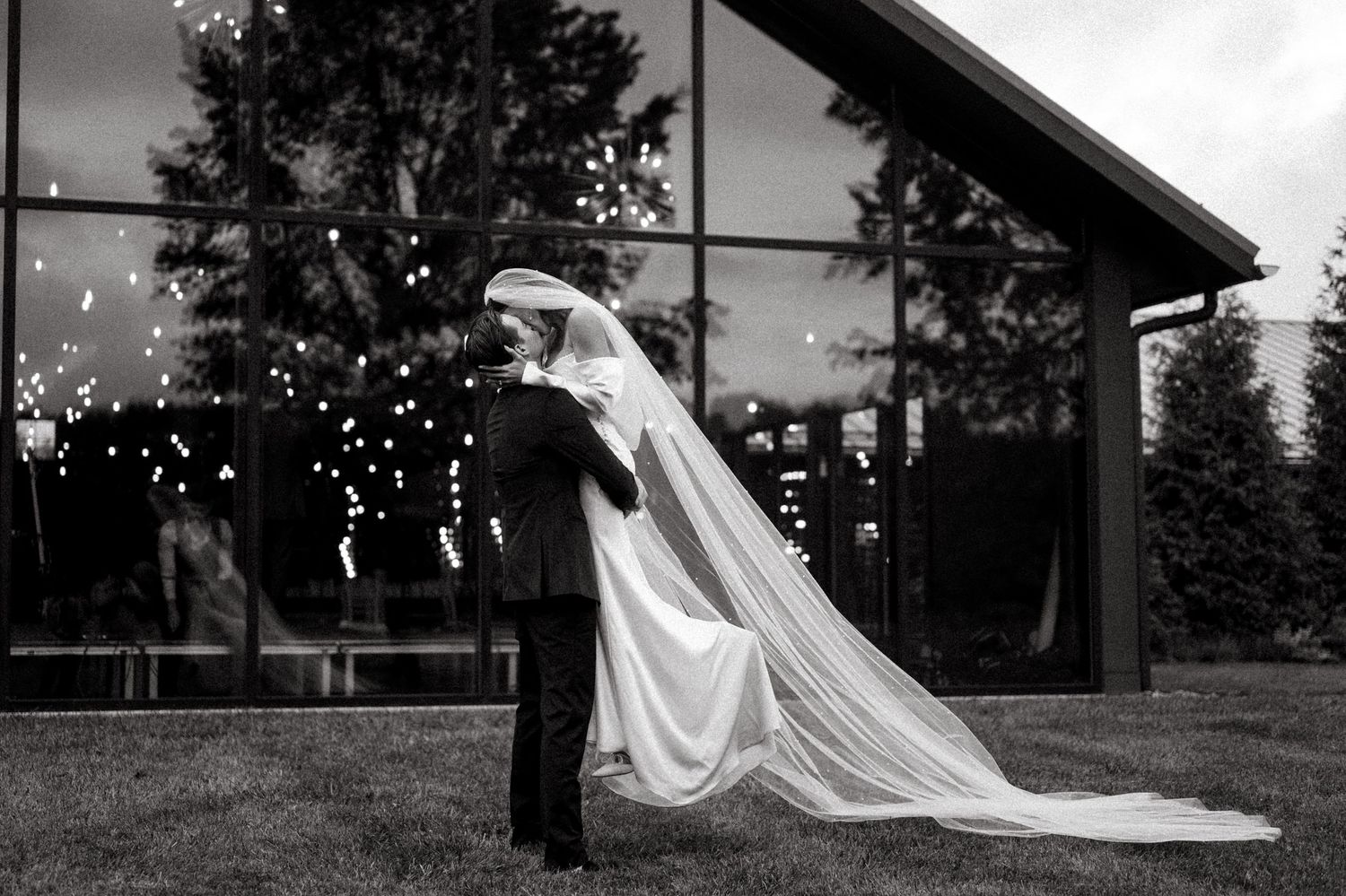 Black and white silhouette of newlyweds kissing outside venue with twinkling lights.