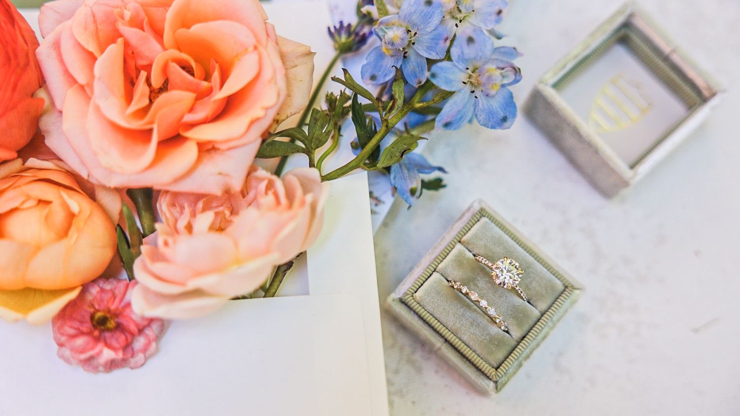 Beautiful peach roses and blue forget-me-nots arranged next to grey velvet ring boxes on a white surface.