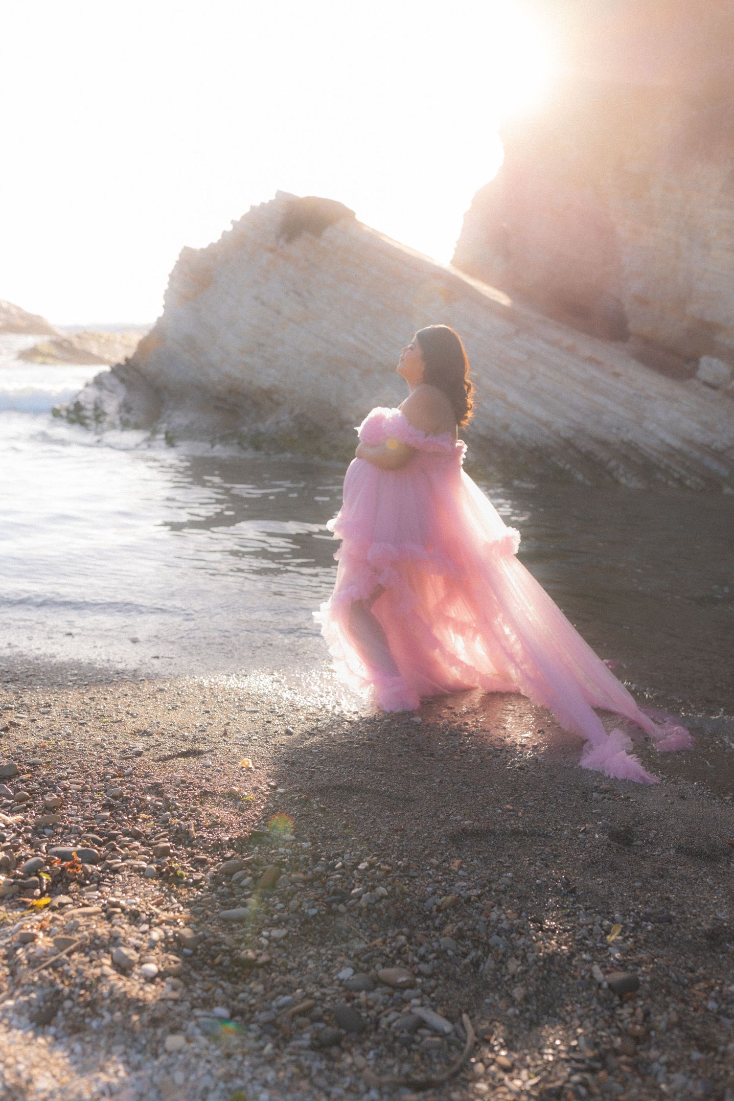 person standing on a rocky beach with coastal cliffs in back, person wearing pink flowy dress in soft warm natural light