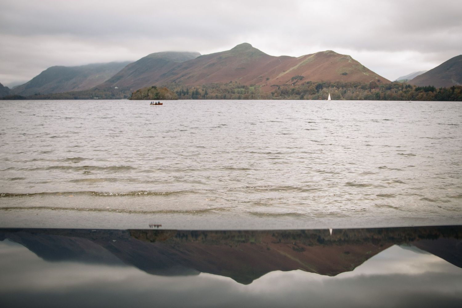 Misty mountains reflect perfectly in the calm waters of a lake with small boats visible in the distance.