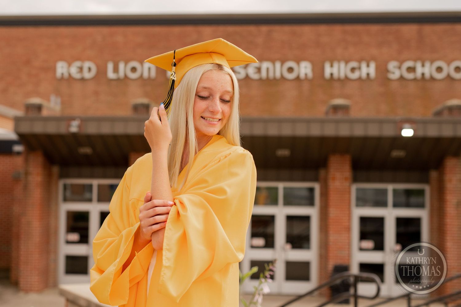 Graduate in yellow cap and gown stands thoughtfully outside brick school building.