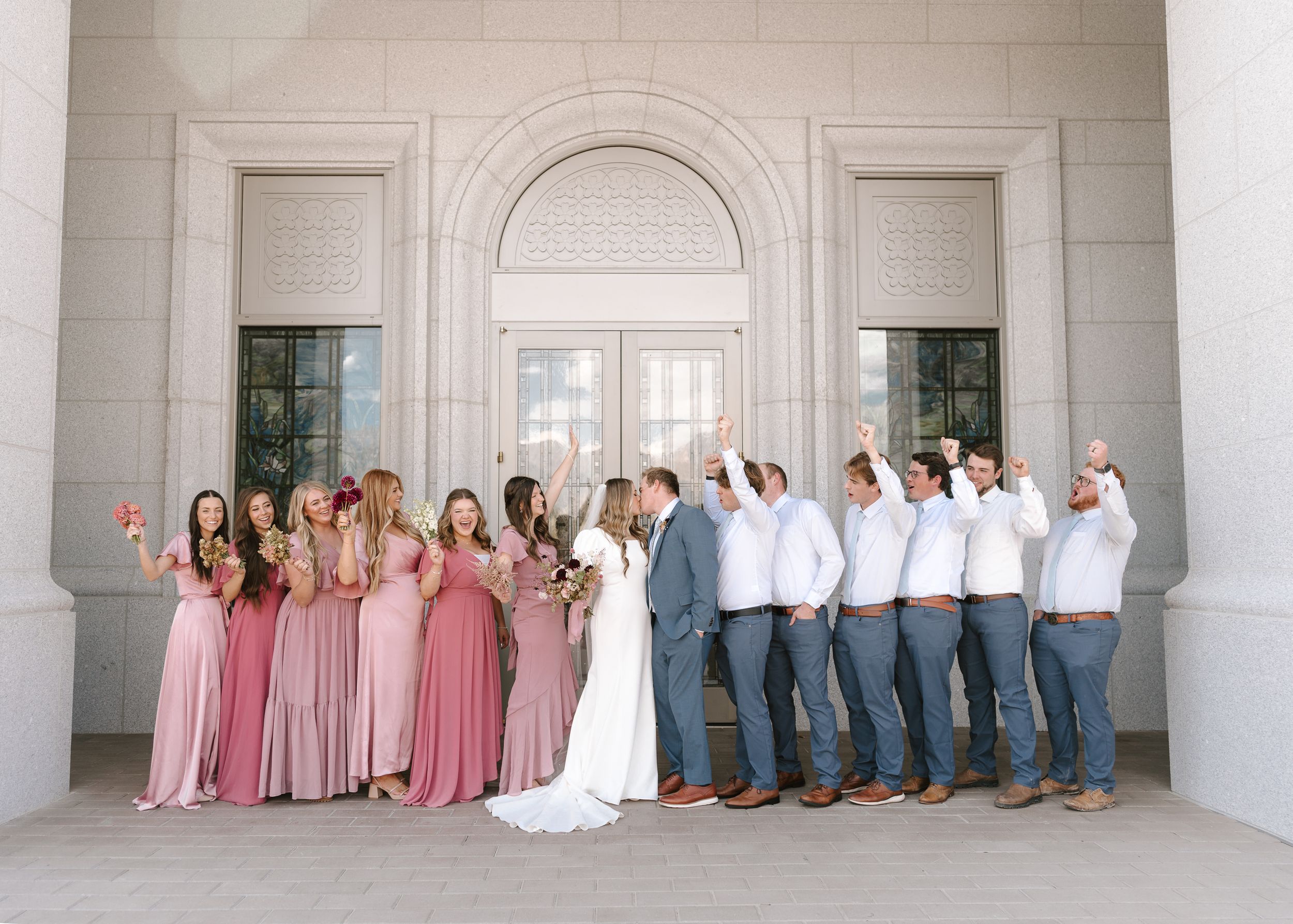 Wedding party in pink dresses and grey suits posing against white stone building.