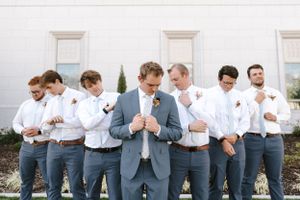Groomsmen in matching gray suits and white shirts gather together while adjusting their ties outside a white building.