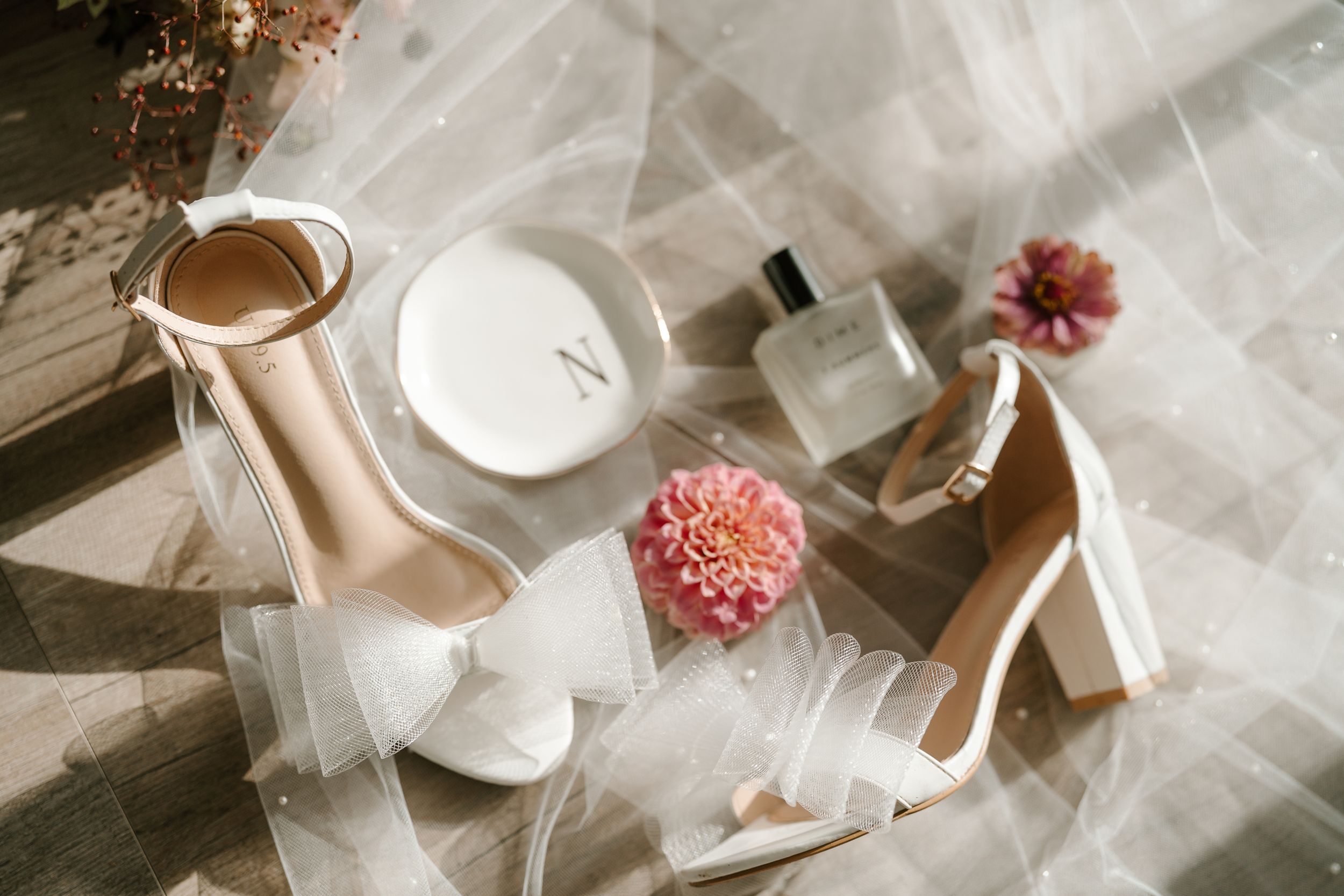 Wedding accessories flatlay featuring white heels, perfume bottle and pink flowers.