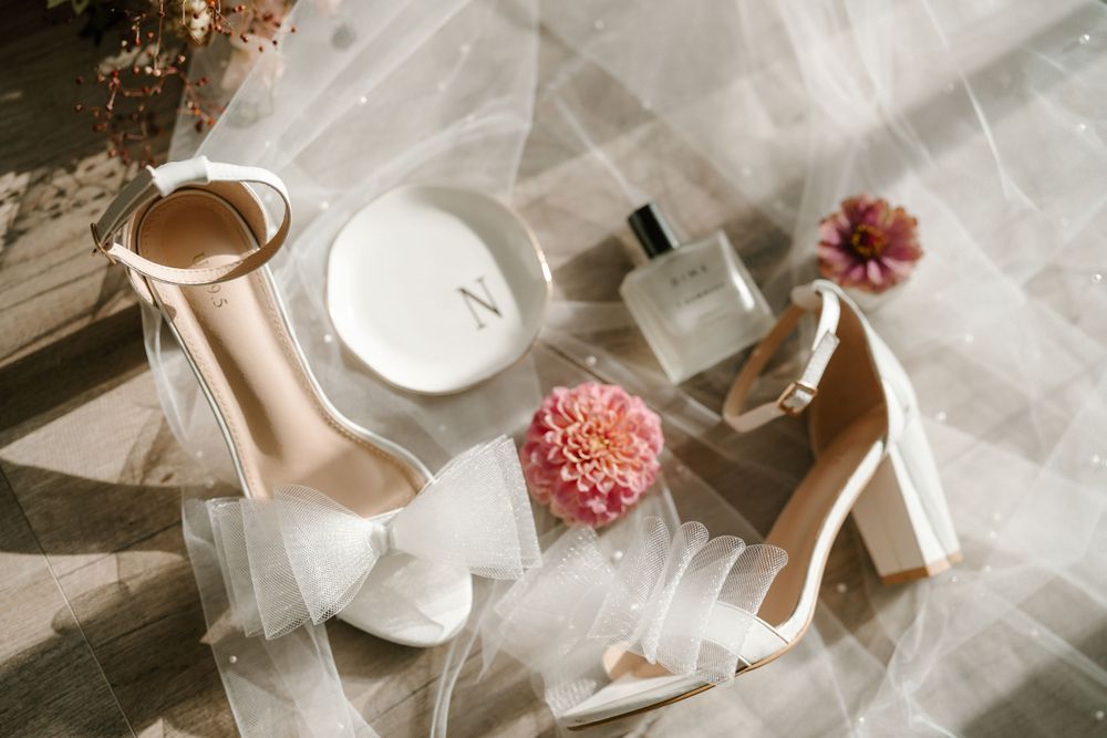 Wedding accessories flatlay featuring white heels, perfume bottle and pink flowers.