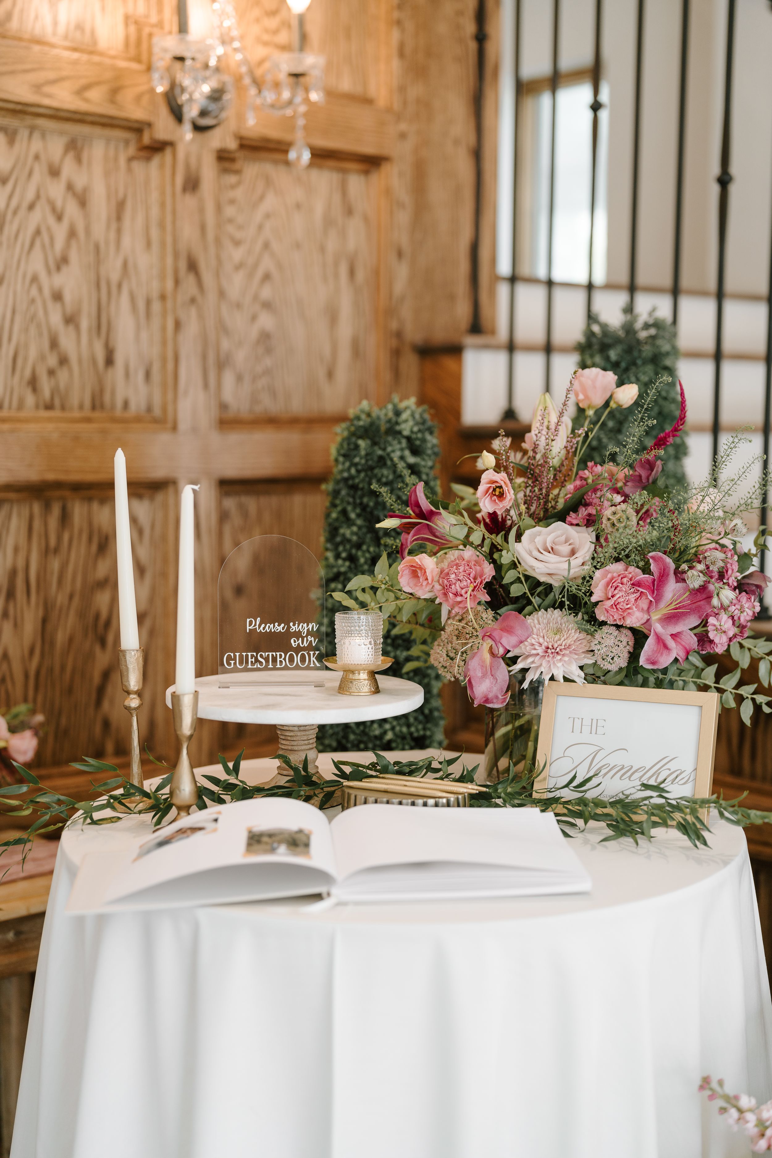 Elegant wedding dessert table with white linens, taper candles, and pink floral arrangements in a rustic wood-paneled venue.