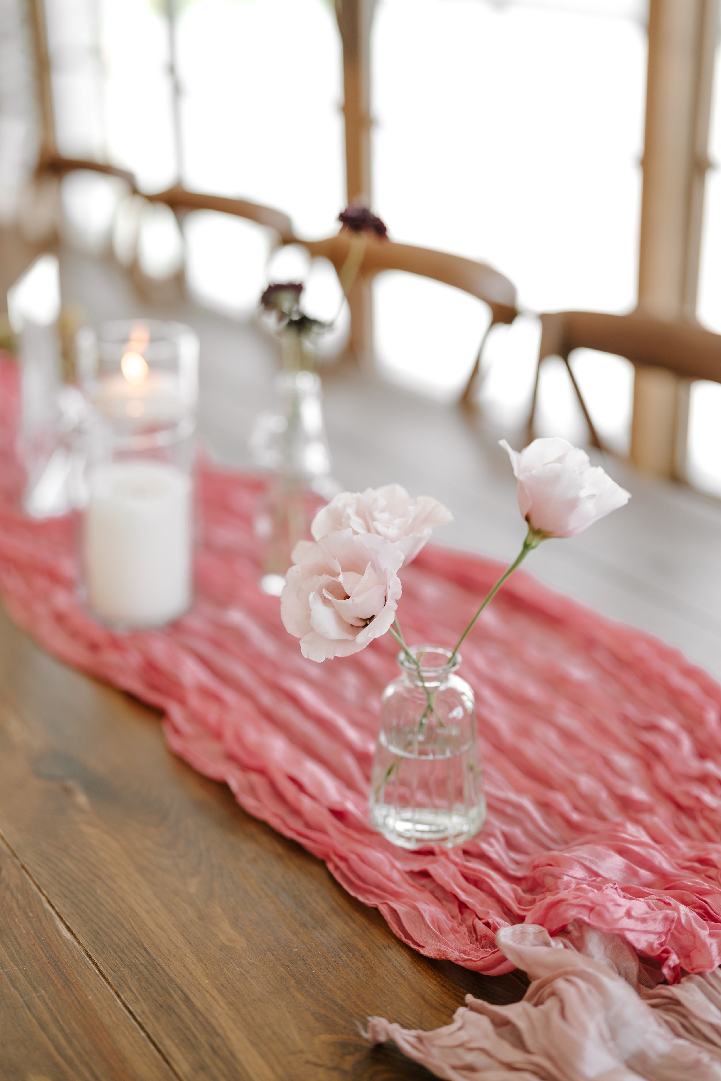 Single pink rose in glass vase on coral table runner with white candles.