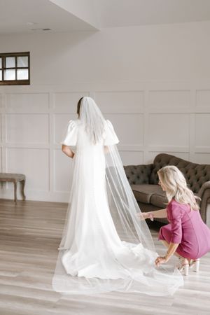 Long white wedding dress with flowing veil being arranged in bright interior room.