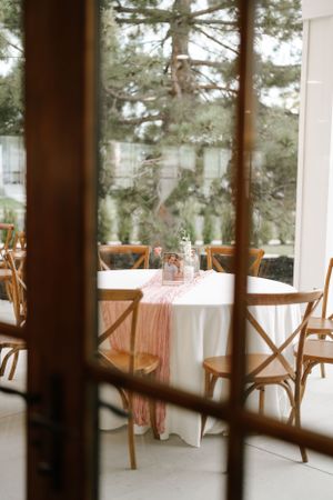 Rustic reception setting with wooden cross-back chairs and pink table accents viewed through window.