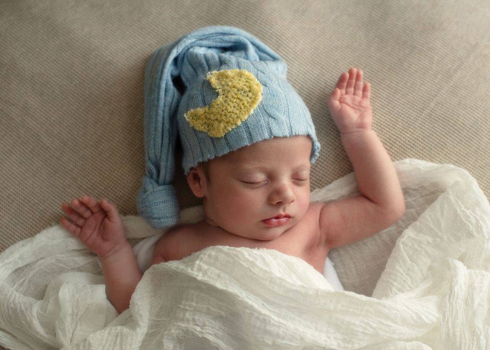 Sleeping infant wearing blue knit cap with gold detail rests peacefully on white blanket.