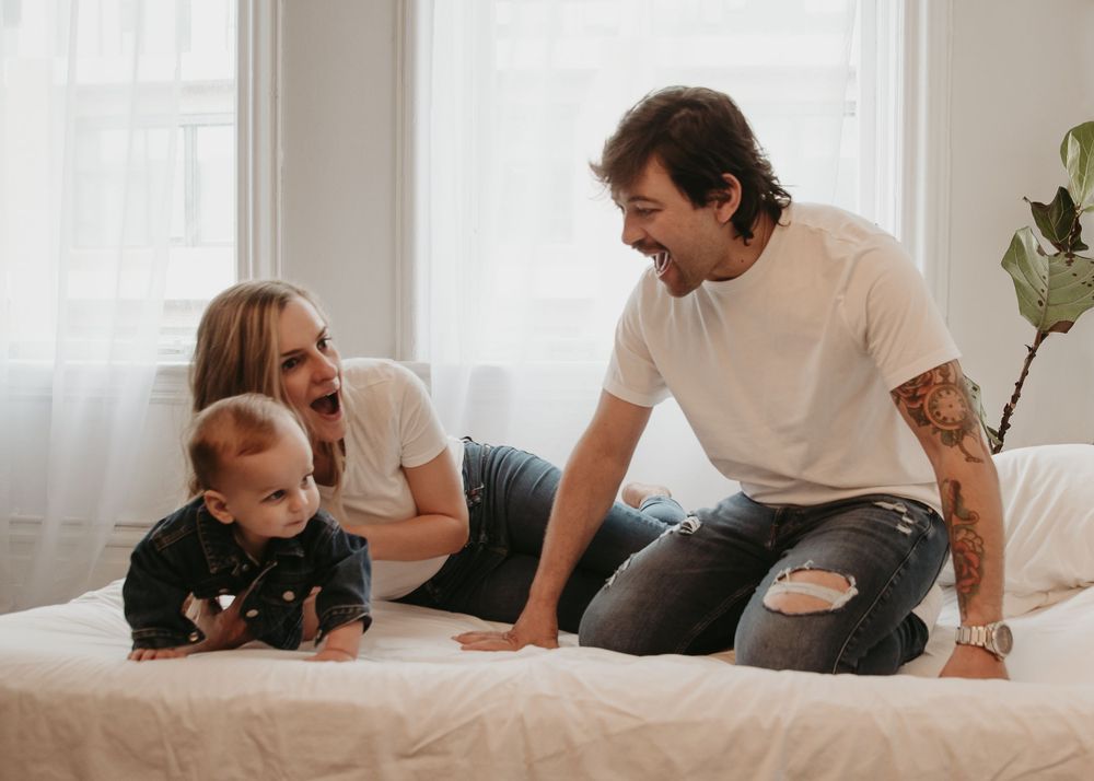 A family sharing playful moments together on a white bed near a window.